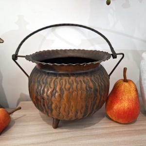 May include: A copper-coloured cauldron-shaped pot with a decorative wavy pattern and a handle. The pot has three small legs and a scalloped rim. A pear with red and yellow hues sits next to the pot.