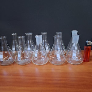 May include: A collection of clear glass Erlenmeyer flasks, with a conical body and a narrow neck, arranged on a wooden surface. A red and black lighter is placed to the right of the flasks. The flasks have white markings.