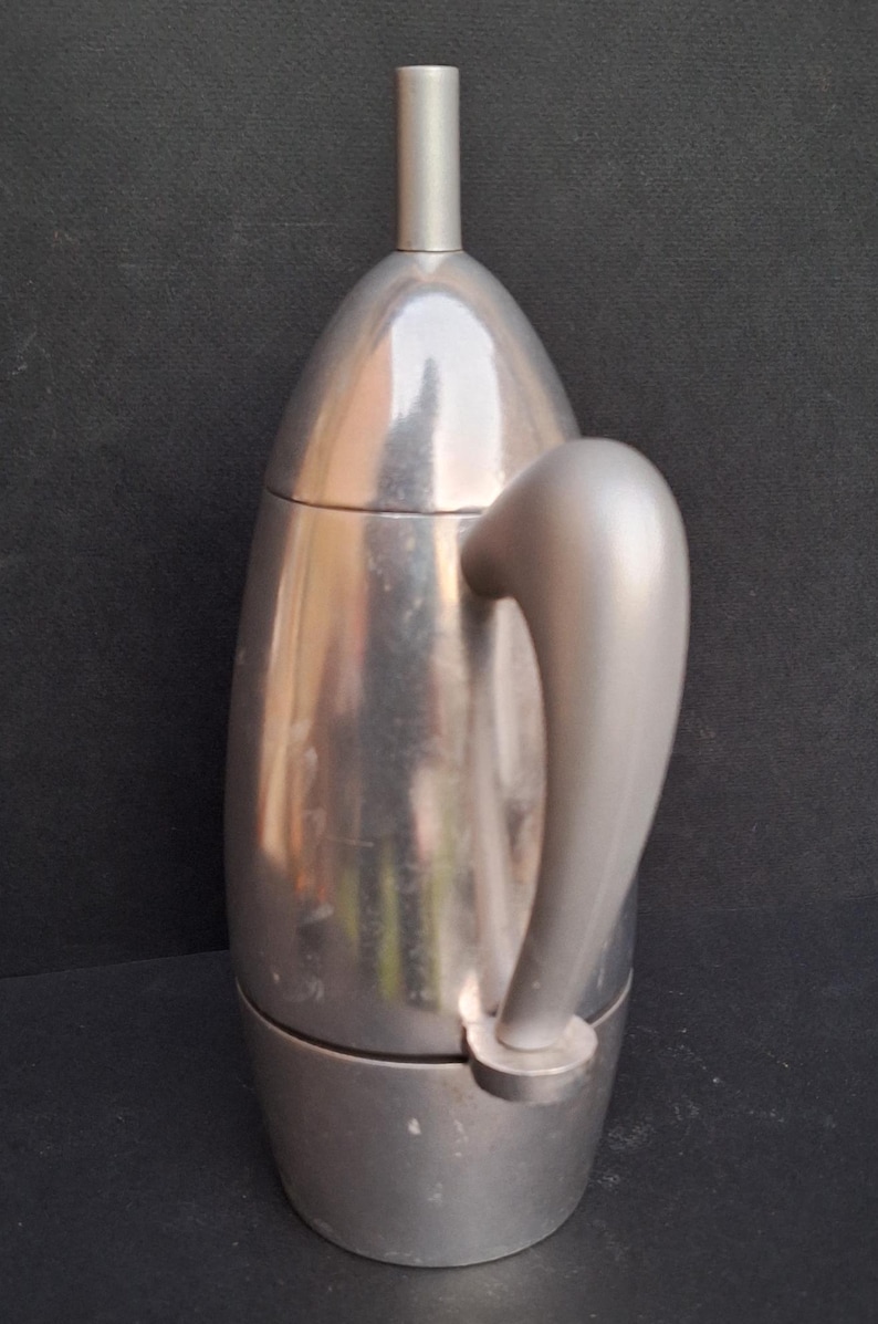 May include: A silver, stovetop espresso maker with a rounded, teardrop-shaped design and a long, curved handle.