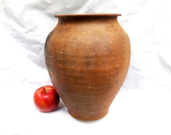 May include: A large, brown, earthenware pot with a rounded body and a wide opening. The pot is sitting on a white surface next to a red apple.