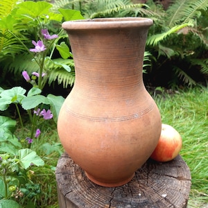 Può includere: Un'anfora di terracotta rustica di colore terracotta con un'ampia apertura e un corpo arrotondato. L'anfora è appoggiata su un ceppo di legno in un giardino.