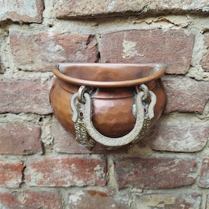 Op de afbeelding: Een halfronde wandplantenbak van gehamerd koper met een gladde, afgeronde rand. De houder is voorzien van een decoratieve, getextureerde metalen handgreep aan de voorzijde, met ingewikkelde gegraveerde patronen en kleine ingelegde details.