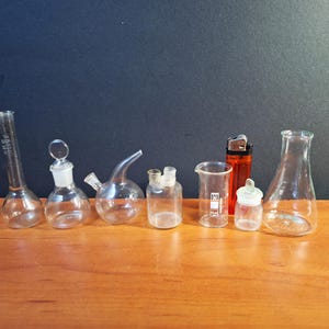 Pode incluir: Uma coleção de béqueres, balões e frascos de laboratório de vidro transparente de várias formas e tamanhos. Um isqueiro laranja também está presente. Os itens estão dispostos em uma superfície de madeira contra um fundo azul escuro.