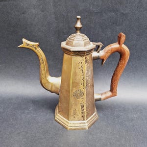 May include: Antique brass coffee pot with a wooden handle. The pot has a unique octagonal shape and a decorative spout. The handle is curved and made of a dark wood. The lid is topped with a small finial. A vintage kitchenware item.