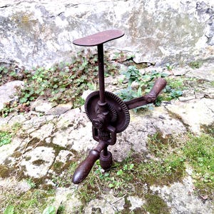 Op de afbeelding: Een vintage schoenmakersstandaard van gietijzer met een verticale paal met een platte ovale bovenplaat, een handmatig aangedreven tandwielmechanisme en een houten handvat. Het metalen oppervlak heeft een donkere, geoxideerde bruine patina.