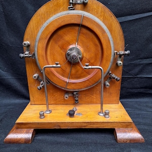 May include: A vintage wooden scientific instrument featuring a large circular wooden disc with a metallic ridged outer edge. The device includes a central rotating metal arm, various silver-toned metal terminals, screws, and a sturdy rectangular wooden base.