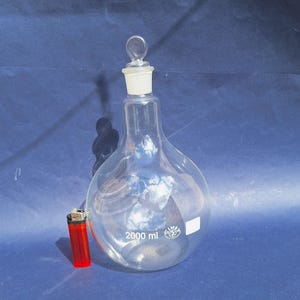 May include: A clear, pear-shaped laboratory flask with a glass stopper. The flask has a capacity of 2000 ml and a white label. A red lighter is next to the flask. The flask is made of clear glass.