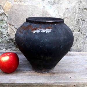 May include: A rustic, black, antique cooking pot with a wide body and a narrow base. The pot has a weathered appearance with areas of rust and wear. A red apple sits next to the pot. The pot is on a wooden surface.