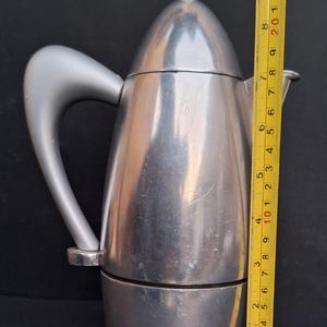 May include: A silver-colored stovetop espresso maker with a curved handle. The maker is 9 inches tall.