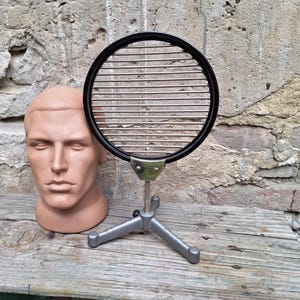 May include: A circular metal grid with a black outer rim, mounted on a silver-coloured adjustable tripod stand. The grid features horizontal parallel bars and is attached to the base with a metal bracket secured by two visible rivets.