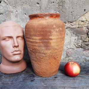 May include: A large, terracotta-coloured clay pot with a wide mouth and a rounded base. The pot has a rustic, aged appearance with visible cracks and imperfections. It is sitting on a wooden surface next to a red apple.