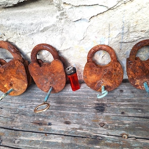 May include: Four old, rusty padlocks with keys, displayed on a weathered wooden surface. The locks are different shapes and sizes, showing significant corrosion. A red lighter is placed between the locks. The background is a textured stone wall.