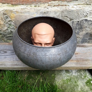 May include: A mannequin head is placed inside a large, round, metal pot. The pot is sitting on a wooden surface.