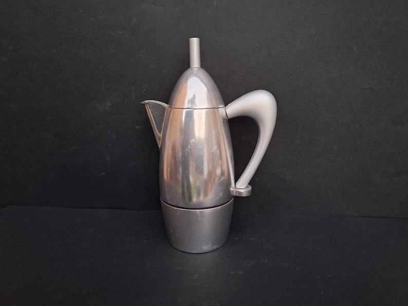 May include: A silver aluminum stovetop espresso maker with a curved handle and a pointed lid.