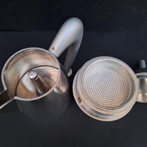 May include: A silver and gray stovetop espresso maker with a removable filter basket. The maker has a handle and a spout for pouring coffee.