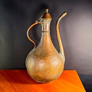 Op de afbeelding: Een hoge, bolvormige koperen kan met een lange, slanke, gebogen tuit en een bijpassend gebogen handvat. Het vat heeft een smalle hals, een klein deksel met een puntige knop en een verweerde, geoxideerde patina op het metalen oppervlak.