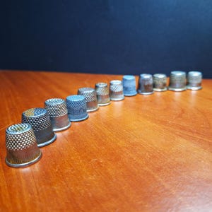 Könnte beinhalten: Eine Reihe von 13 silbernen Fingerhüten, die auf einer Holzoberfläche aufgereiht sind. Die Fingerhüte sind in einem leichten Bogen angeordnet, wobei der größte Fingerhut vorne und der kleinste Fingerhut hinten liegt.