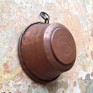 May include: A round copper bowl with a weathered, metallic finish and a dark patina. The bowl features a circular base and a small, integrated metal ring attached to the top rim, allowing it to be hung vertically against a textured surface.