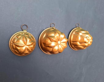 Puede incluir: Tres moldes de metal de color cobre con forma circular y un patrón floral en relieve en la superficie. Cada molde cuenta con una pequeña anilla de metal unida al borde superior, lo que permite colgarlos. Tienen un acabado metálico brillante.