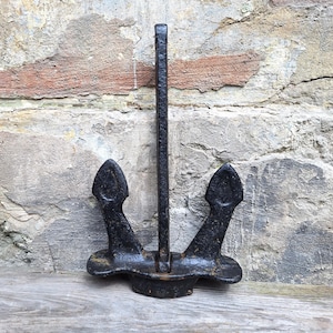 Könnte beinhalten: Ein schwarzer Metallanker mit verwittertem Aussehen. Der Anker hat zwei Flunken und einen langen Schaft und ruht auf einer Holzoberfläche vor einer strukturierten Backsteinmauer. Der Anker ist wahrscheinlich ein dekoratives nautisches Objekt.