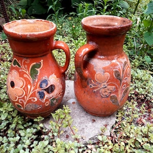 May include: Two terracotta pitchers with a rounded body, a wide flared rim, and a single side handle. Each vessel features hand-painted floral and butterfly motifs in muted tones of green, dark blue, and cream against the reddish-brown clay surface.