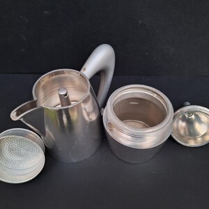 May include: A silver aluminum stovetop espresso maker with a removable filter basket and a separate lid. The maker is disassembled, showing the interior of the pot and the filter basket.