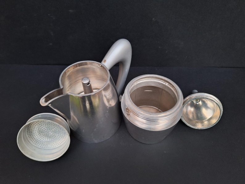 May include: A silver aluminum stovetop espresso maker with a removable filter basket and a separate lid. The maker is disassembled, showing the interior of the pot and the filter basket.