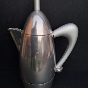 May include: A silver-colored stovetop espresso maker with a rounded, modern design. The maker has a removable top and a handle that curves upwards.