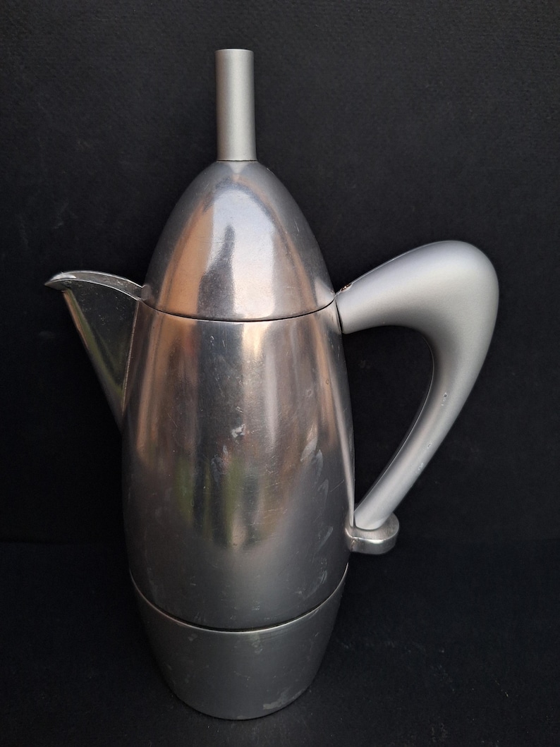 May include: A silver-colored stovetop espresso maker with a rounded, modern design. The maker has a removable top and a handle that curves upwards.