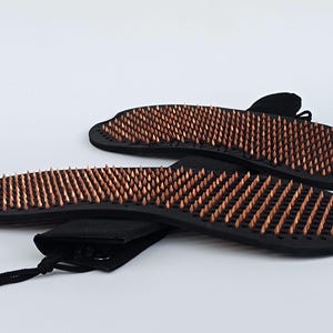 Op de afbeelding: Twee zwarte sadhu-planken met koperkleurige spijkers. De planken zijn ovaal van vorm en hebben talrijke spijkers die uit het oppervlak steken. Een zwart trekkoordzakje is inbegrepen.