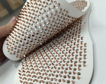 Puede incluir: Esterilla de masaje blanca con numerosas púas de color cobre. La esterilla tiene un diseño perforado con agujeros espaciados uniformemente. Las púas están dispuestas en un patrón de cuadrícula, creando una superficie texturizada. La esterilla está diseñada para uso terapéutico.