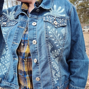 May include: A blue denim jacket with white painted floral designs and silver studs. The jacket has a button closure and a plaid shirt underneath.