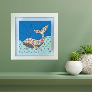 May include: A square white frame containing a layered fabric art piece. The artwork features a whale with a floral pattern on a blue background, positioned above a light blue section decorated with small pink and green floral motifs.