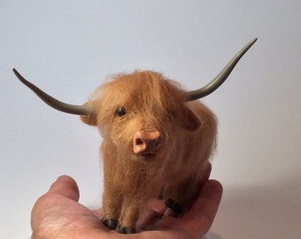 Op de afbeelding: Een klein, bruin, pluche Highland-koe-beeldje met grijze hoorns. Het beeldje zit op de hand van een persoon.