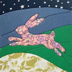 May include: A fabric collage featuring a leaping rabbit made from pink floral patterned textile. The background consists of layered fabric sections in dark blue with white stars, a muted blue horizon, green hills, and a cream paisley base.
