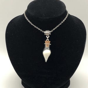 May include: A silver chain necklace with a small glass vial pendant. The vial is filled with white crystals and has a cork stopper.