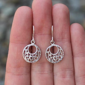 Puede incluir: Un par de pendientes de filigrana plateada. Cada pendiente presenta un diseño circular con un patrón calado de líneas onduladas y detalles en forma de corazón. Los pendientes tienen un cierre de gancho sencillo.