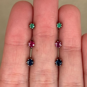 May include: A pair of vertical drop earrings, each featuring three round-cut gemstones set in silver-toned metal. The stones are arranged in a column, with a green gem at the top, a pink gem in the center, and a blue gem at the bottom of each earring.