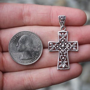 May include: A silver pendant in the shape of a cross with an ornate, openwork design. The cross is held next to a U.S. quarter for size comparison. The cross features floral and heart-shaped details. The pendant is likely made of sterling silver.