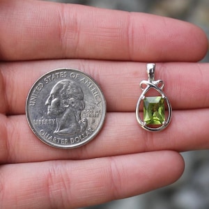 May include: A silver pendant with a rectangular, light green gemstone. The pendant is held next to a U.S. quarter for size comparison. The gemstone is approximately 0.25 inches in length.