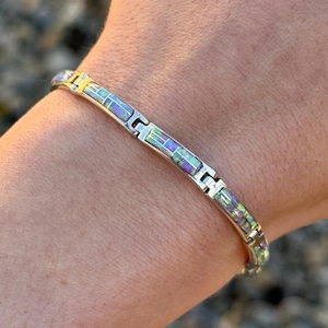 以下が含まれることがあります： 長方形の虹色の象嵌が施されたシルバーブレスレット。ブレスレットはシルバー製で、それぞれが光沢のあるカラフルな石で象嵌された一連の長方形のセグメントが特徴です。ブレスレットは手首に着用されています。