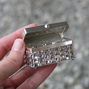 Könnte beinhalten: Eine kleine silberne Metallbox mit einem gewebten Muster auf dem Boden und einem Scharnierdeckel. Die Box ist offen und scheint leer zu sein.