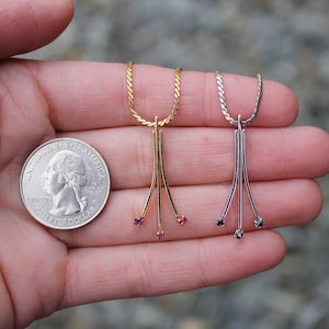 May include: Two delicate necklaces with three small gemstones at the end of each pendant. The necklace on the left has a gold chain and pink gemstones. The necklace on the right has a silver chain and black gemstones. A US quarter is shown for scale.