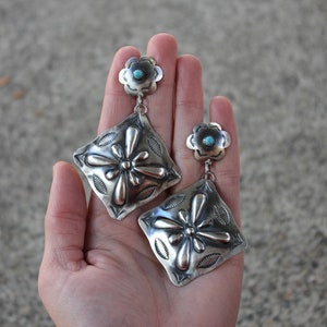 Könnte beinhalten: Ein Paar silberne Ohrringe mit einem Türkis-Stein in der Mitte jeder blütenförmigen Oberseite. Die Ohrringe haben ein quadratisches Design mit einem Blumenmuster.