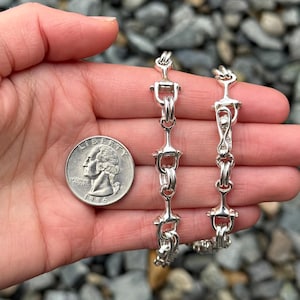 May include: Two silver bracelets with a distinctive equestrian design are shown. The bracelets are held in a hand next to a quarter coin. The bracelets feature a series of linked, stylized horse bit shapes.
