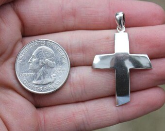 May include: A silver cross pendant, shown next to a US quarter for size comparison. The cross is simple and has a slightly rounded shape.
