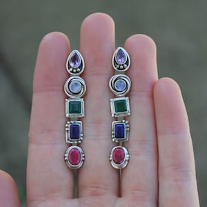 May include: A pair of silver earrings with a cascade of gemstones. The earrings feature a teardrop-shaped amethyst, a round moonstone, a square green stone, a rectangular blue stone, and an oval pink stone.