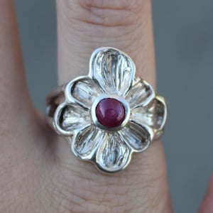 May include: A silver ring featuring a flower design with a red gemstone at its centre.