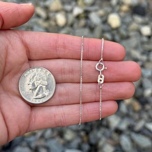 May include: A silver box chain necklace with a lobster clasp is held in a hand next to a quarter. The chain is delicate and has a square link design. The quarter is dated 1996.