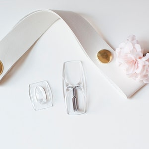 May include: White leather strap with a gold metal eyelet, two clear plastic adhesive hooks, and a pink fabric flower.
